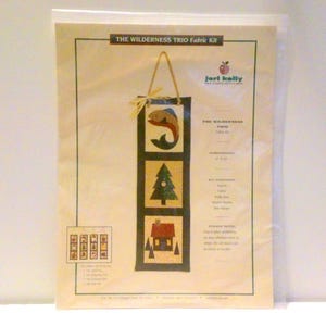 Può includere: Un kit di tessuto per un'appendino da parete intitolato "The Wilderness Trio" di Jert Kelly. Il kit include tessuto, ovatta, filo e un gancio. Il design presenta un pesce, un albero e una casa.