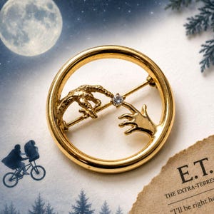 May include: A circular gold-toned brooch featuring two sculpted hands reaching toward each other, with a single clear round gemstone set between the fingertips. The design is inspired by a cinematic scene, with a pin mechanism on the back.