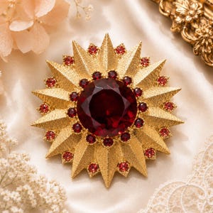 May include: A gold-toned starburst brooch with a large, round, deep red center stone and smaller red accent stones. The brooch has a textured surface and a sunburst design, creating a striking and elegant accessory.