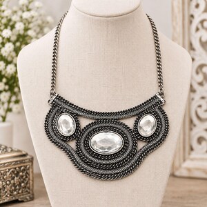 May include: A silver-toned statement necklace featuring a thick chain design with three large, clear, oval-shaped faceted stones. The metalwork consists of intricate, textured, dark silver links arranged in concentric patterns around the stones.