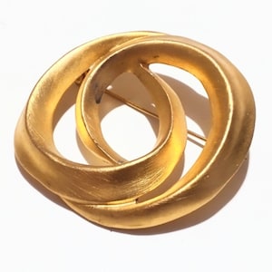 May include: A gold-toned brooch with a double-loop design. The brooch has a pin on the back for secure fastening. The metal has a brushed texture, and the overall shape is circular. The brooch is set against a white background.