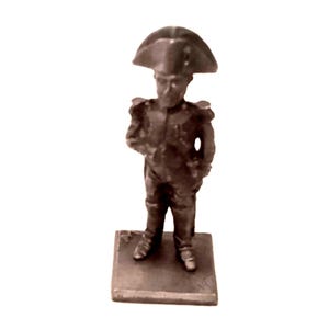 May include: A small, metallic figurine of a historical figure, likely Napoleon Bonaparte, in a military uniform and iconic hat. The figure stands on a rectangular base, with details visible in the uniform and facial features. The figurine is a muted bronze color.