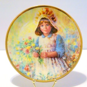 May include: A porcelain plate with a gold rim features a painting of a young girl in a white dress and blue jacket holding a bouquet of flowers. The background is a soft yellow and green with a watercolour effect. The plate is signed "De  Wilde" in the lower right corner.