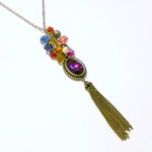 May include: A vintage-style necklace with a gold-tone chain. The pendant features a large, oval, purple gemstone surrounded by a decorative frame. A cluster of colorful beads and a gold tassel hang below the gemstone, creating a unique and eye-catching design.