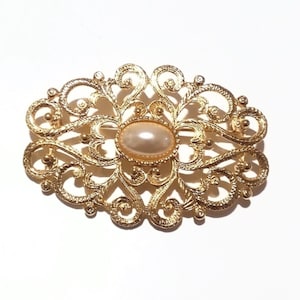 May include: Gold-toned filigree brooch with an oval, cream-colored center stone. The brooch features an ornate, openwork design with swirling patterns and small, round accents. A classic jewelry piece.