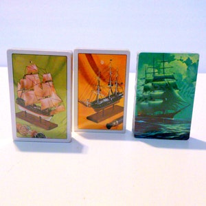 May include: Three playing cards featuring illustrations of sailing ships. The cards have white borders and varying background colors: green, orange, and teal. The ships are depicted with detailed rigging and sails, mounted on wooden bases.