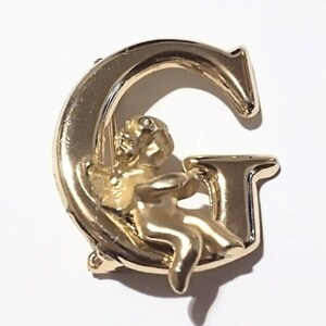 May include: A gold-colored metal brooch in the shape of the letter "G". A small cherub figure is seated within the curve of the letter. The brooch has a shiny, polished finish and is set against a white background.
