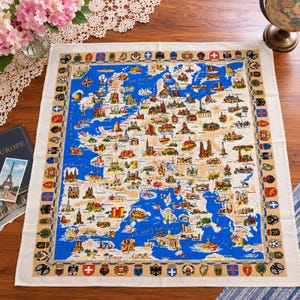 May include: A rectangular textile featuring a colorful map of Europe with blue seas and beige landmasses. The map displays various landmarks, cities, and ships, surrounded by a decorative border of national crests and flags on a tan background.