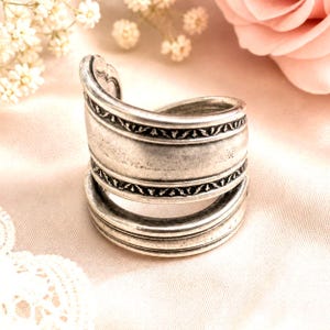 May include: A silver-toned ring with an intricate design. The ring appears to be made from repurposed silverware, featuring decorative bands with a repeating pattern. The ring is displayed on a soft, pink fabric, with flowers in the background.