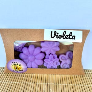 Puede incluir: Una caja de cartón marrón con forma de almohada y una ventana rectangular transparente que muestra cuatro ceras aromáticas en forma de flor de color violeta claro. Una etiqueta blanca dice Violeta y una pegatina ovalada morada muestra un gato.