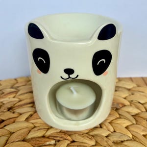 Puede incluir: Un calentador de cera de cerámica color crema con forma de cara de panda, con orejas negras, manchas negras en los ojos, una nariz pequeña y negra y una boca sonriente. Una vela de té blanca se encuentra dentro de la abertura circular.