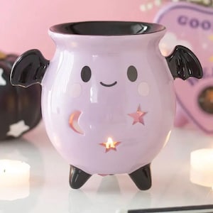 May include: A light purple ceramic wax warmer shaped like a cauldron with black bat wings and three black feet. The front features a smiling face, a crescent moon cutout, and star cutouts with a glowing light visible from within the vessel.