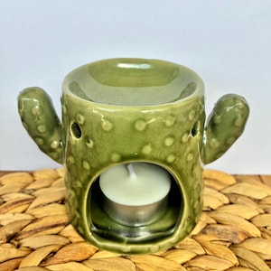 Puede incluir: Un quemador de aceite de cerámica verde salvia con forma de cactus y superficie texturizada con pequeños puntos en relieve. El cuerpo cilíndrico tiene dos brazos laterales, un cuenco superior para aceite y una abertura frontal.