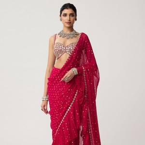 May include: A striking red saree with a sequined top and matching draped fabric. The saree has a ruffled edge and is decorated with small, scattered embellishments. A matching red train extends from the back.