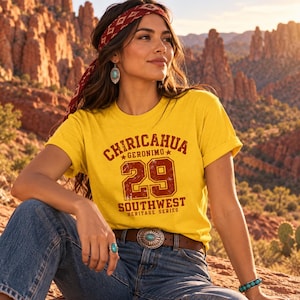 Può includere: Una t-shirt gialla a maniche corte con testo bordeaux dall'effetto consumato che recita CHIRICAHUA GERONIMO 29 SOUTHWEST HERITAGE SERIES. La maglia è abbinata a jeans blu e a una cintura in pelle marrone con una grande fibbia in argento e turchese.