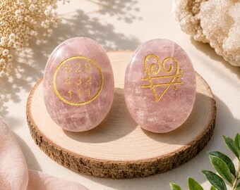 Puede incluir: Dos piedras de cuarzo rosa lisas de forma ovalada con grabados de color dorado. La piedra de la izquierda muestra un círculo con los números 528, 432 y 111. La piedra de la derecha presenta un símbolo de corazón dorado con base triangular.