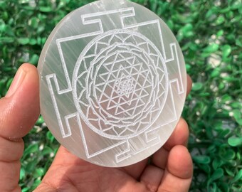 May include: A round, white selenite crystal with an etched Sri Yantra design. The crystal is held in a hand against a backdrop of green foliage. The intricate geometric pattern is a central focus.