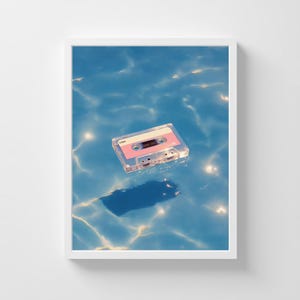 May include: A framed print featuring a vintage cassette tape floating on a blue water surface. The cassette has a pink label and a clear plastic casing. The water has light reflections, creating a serene and nostalgic scene.