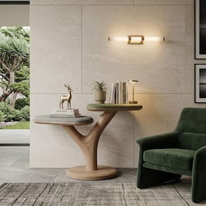 May include: A two-tiered side table with a light wood base shaped like a tree trunk, featuring two oval platforms with grey and green upholstered tops. The table holds a gold deer figurine, a potted plant, several books, and a small lamp.