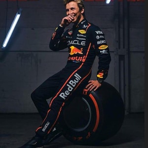 Może przedstawiać: Osoba w czarno-pomarańczowym kombinezonie wyścigowym Red Bull z logo sponsor&oacute;w i pomarańczowymi paskami, oparta o dużą oponę. Osoba się uśmiecha.