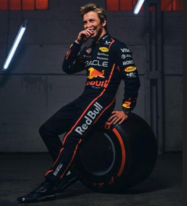 Może przedstawiać: Osoba w czarno-pomarańczowym kombinezonie wyścigowym Red Bull z logo sponsor&oacute;w i pomarańczowymi paskami, oparta o dużą oponę. Osoba się uśmiecha.