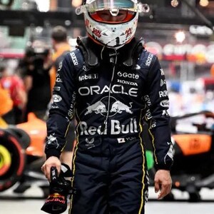 Może przedstawiać: Kierowca Formuły 1 w granatowym kombinezonie wyścigowym z ż&oacute;łtymi akcentami i białym kasku z czerwonymi i czarnymi detalami. Kombinezon posiada logo sponsor&oacute;w, takich jak "Oracle" i "Red Bull". Kierowca trzyma czarny but wyścigowy.