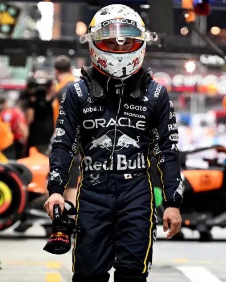 Może przedstawiać: Kierowca Formuły 1 w granatowym kombinezonie wyścigowym z ż&oacute;łtymi akcentami i białym kasku z czerwonymi i czarnymi detalami. Kombinezon posiada logo sponsor&oacute;w, takich jak "Oracle" i "Red Bull". Kierowca trzyma czarny but wyścigowy.