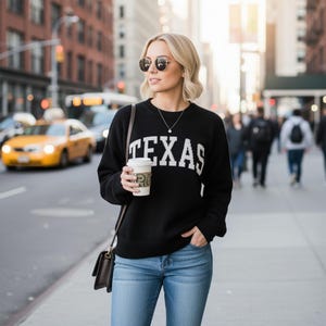 May include: A black long-sleeve knit sweater featuring the word TEXAS in white block lettering across the chest, paired with blue denim jeans, a dark brown shoulder bag, a silver necklace, and round-frame sunglasses while holding a coffee cup.