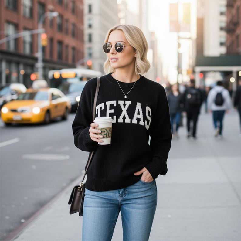 May include: A black long-sleeve knit sweater featuring the word TEXAS in white block lettering across the chest, paired with blue denim jeans, a dark brown shoulder bag, a silver necklace, and round-frame sunglasses while holding a coffee cup.