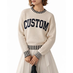 May include: A cream-colored long-sleeve knit sweater featuring the word CUSTOM in dark navy blue block lettering across the chest. The garment includes a ribbed crew neckline, cuffs, and hem, all detailed with a dark navy blue and cream stripe pattern.