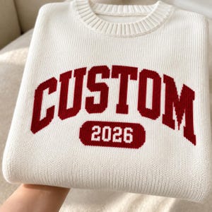May include: A white crew neck sweater featuring the word CUSTOM in large, bold, dark red block letters across the chest, with the number 2026 printed in white inside a dark red horizontal oval shape positioned directly below the text.