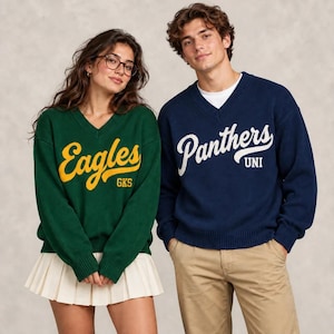 May include: A dark green V-neck sweater with yellow text reading Eagles GKS and a navy blue V-neck sweater with white text reading Panthers UNI. Both sweaters feature long sleeves with ribbed cuffs and a ribbed V-neckline design.