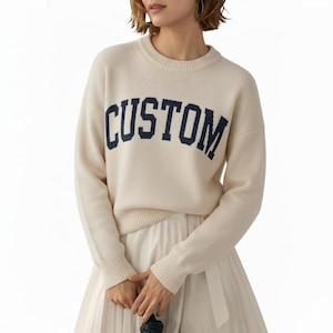 May include: Cream-colored sweater with the word "CUSTOM" in navy blue block letters across the chest. The sweater has a ribbed crew neck, cuffs, and hem. The model is wearing a pleated skirt.