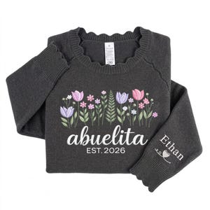 Peut inclure: Pull-over gris fonc&eacute; avec broderie florale rose, violette et blanche. Le mot "abuelita" est brod&eacute; en blanc, avec "EST. 2026" en dessous. Le poignet de la manche porte le nom "Ethan" et un motif de c&oelig;ur.