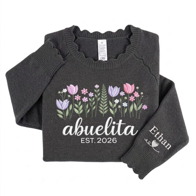 Peut inclure: Pull-over gris fonc&eacute; avec broderie florale rose, violette et blanche. Le mot "abuelita" est brod&eacute; en blanc, avec "EST. 2026" en dessous. Le poignet de la manche porte le nom "Ethan" et un motif de c&oelig;ur.