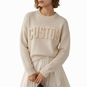 May include: A cream-colored long-sleeve sweater featuring a crew neckline and ribbed cuffs. The word CUSTOM is displayed across the chest in raised, textured, cream-colored knit lettering. The sweater is paired with a white pleated skirt.