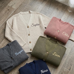 May include: Five folded, personalized cardigans in various colors: beige, dusty rose, dark gray, olive green, and navy blue. Each sweater has a V-neck and button closure, with a name embroidered on the front: Sophia, Chloe, Harper, Liam, and Nora.