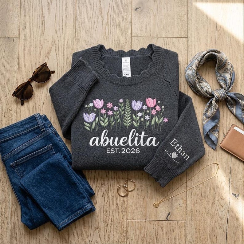Peut inclure: Un pull gris anthracite orn&eacute; de broderies florales roses, violettes et blanches. Le mot "abuelita" est brod&eacute; sous les fleurs, avec "EST. 2026" en dessous. La manche porte le nom "Ethan". Le pull est accompagn&eacute; d'un jean et d'accessoires.
