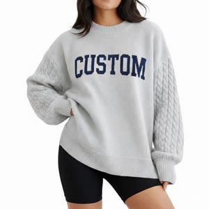 May include: A light gray long-sleeve sweater featuring a crew neckline and ribbed cuffs and hem. The front displays the word CUSTOM in dark blue block lettering. The sleeves are designed with a prominent vertical cable knit texture pattern.