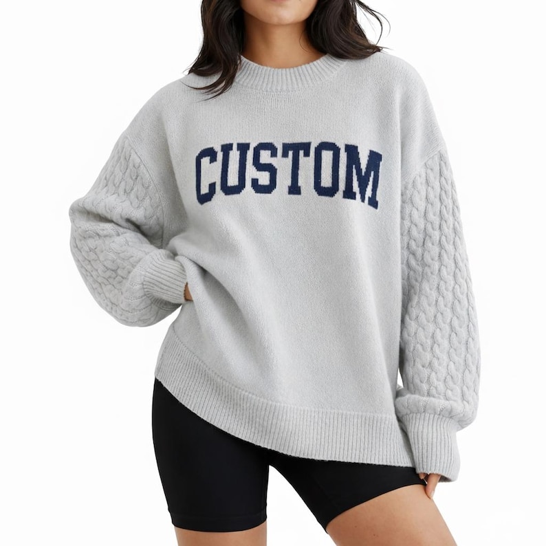 May include: A light gray long-sleeve sweater featuring a crew neckline and ribbed cuffs and hem. The front displays the word CUSTOM in dark blue block lettering. The sleeves are designed with a prominent vertical cable knit texture pattern.