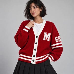 May include: A red V-neck cardigan with white trim, featuring a white letter M on the left chest and the number 68 on the left sleeve. The cardigan has a button-down front, long sleeves with white stripes at the cuffs, and a white hem with stripes.