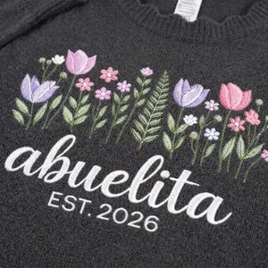 Peut inclure: Pull-over gris fonc&eacute; avec broderie florale rose, violette et blanche. Le mot "abuelita" est brod&eacute; en blanc, avec "EST. 2026" en dessous. Le pull a un col rond.