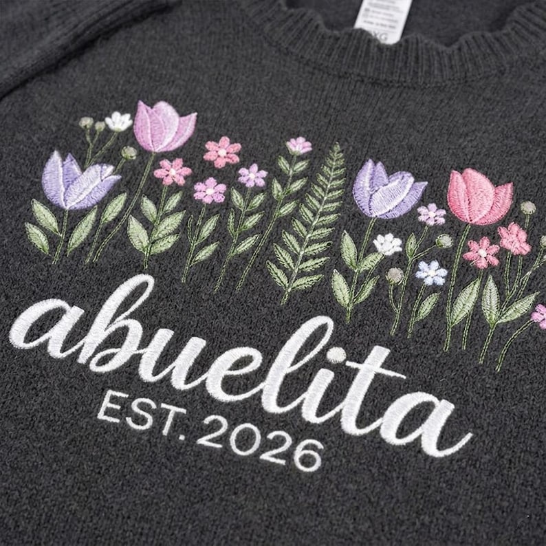 Peut inclure: Pull-over gris fonc&eacute; avec broderie florale rose, violette et blanche. Le mot "abuelita" est brod&eacute; en blanc, avec "EST. 2026" en dessous. Le pull a un col rond.