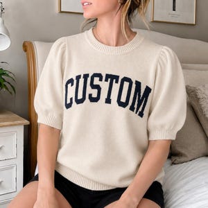 May include: Cream-colored sweater with short, puffed sleeves. The word "CUSTOM" is displayed in navy blue block letters across the chest. The sweater is a casual, comfortable style.