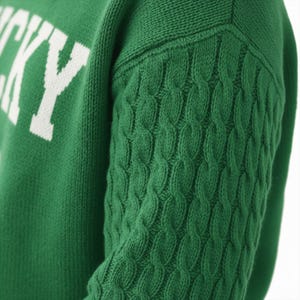 May include: A green knit sweater featuring a cable knit pattern on the sleeve and the white text "LUCKY" printed on the chest. The garment is constructed with a textured knit fabric, showcasing a distinct contrast between the cable knit sleeve.