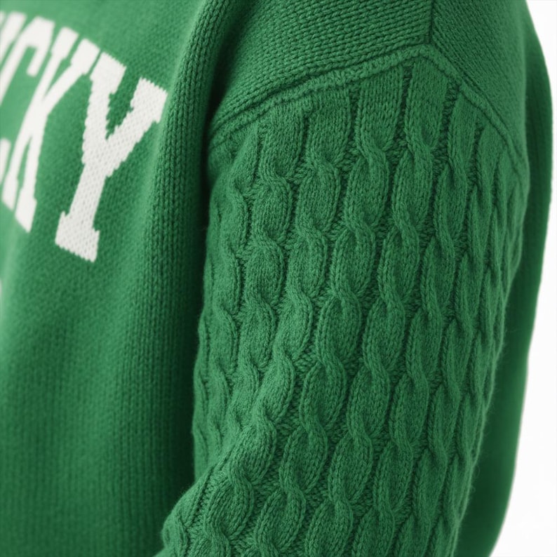 May include: A green knit sweater featuring a cable knit pattern on the sleeve and the white text "LUCKY" printed on the chest. The garment is constructed with a textured knit fabric, showcasing a distinct contrast between the cable knit sleeve.
