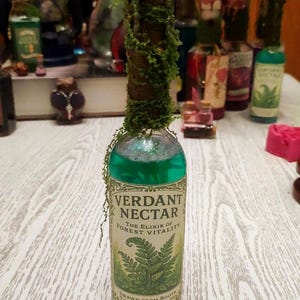 Puede incluir: Una botella de vidrio decorativa llena de líquido verde, etiquetada como "Verdant Nectar: The Elixir of Forest Vitality". La botella está adornada con musgo y una envoltura marrón. Otras botellas son visibles en el fondo.