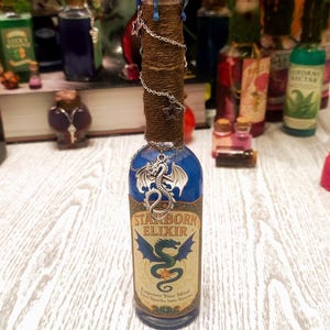 Może przedstawiać: Dekoracyjna szklana butelka z niebieskim i brązowym wzorem. Butelka jest oznaczona napisem "STARBORN ELIXIR" z ilustracją smoka. Ma niebieską pieczęć woskową i jest ozdobiona łańcuszkiem i zawieszkami w kształcie gwiazd. Inne butelki są w tle.