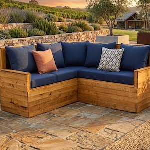May include: A wooden corner sofa with navy blue cushions and accent pillows in orange and white patterns. The sofa is made of light brown wood and is placed on a stone patio. The background features a vineyard and a house.