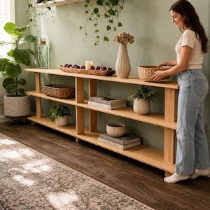Op de afbeelding: Een licht houten boekenplank met drie niveaus, met diverse decoratieve items zoals planten, boeken en een gevlochten mand. De planken worden ondersteund door verticale houten balken. Een vrouw plaatst een mand op de bovenste plank.
